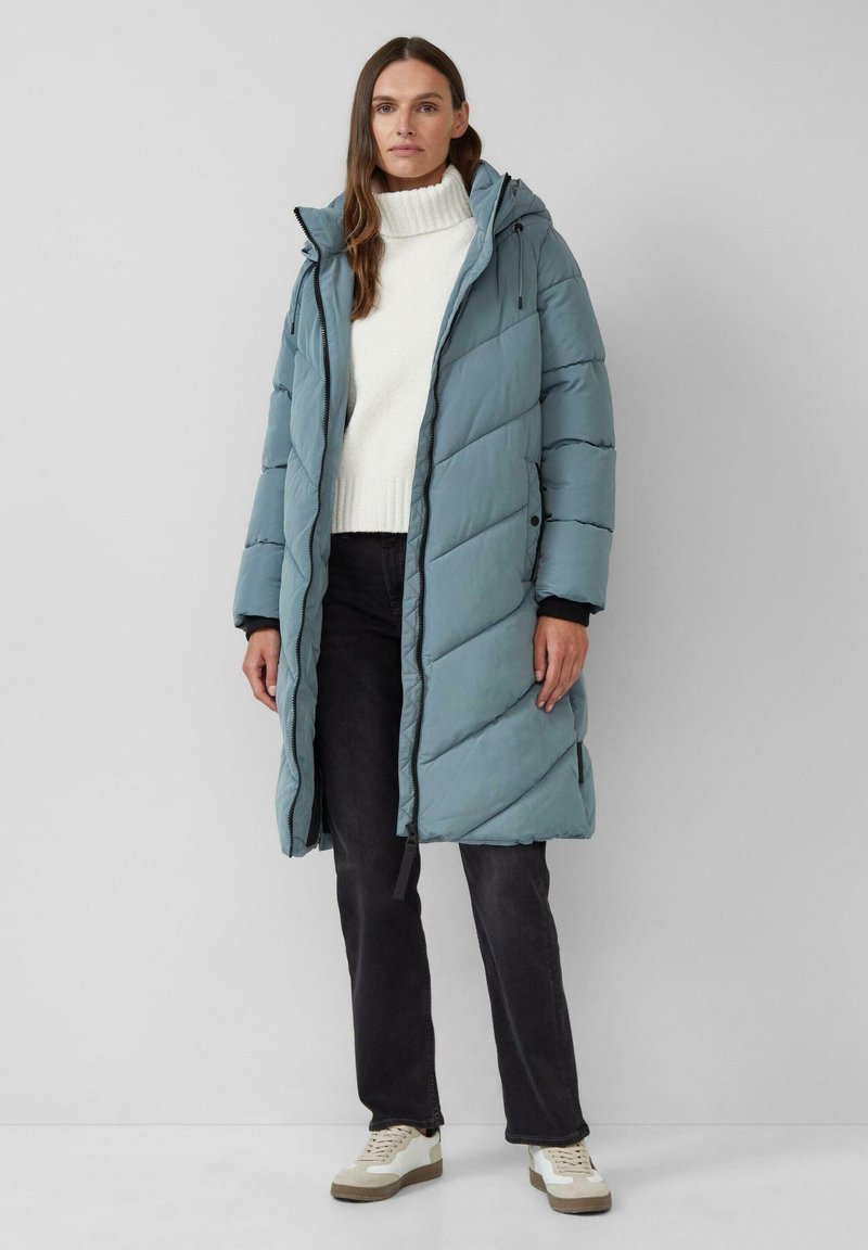 Puffermantel in sanftem Blau mit diagonaler Steppung, Kapuze und seitlichen Reißverschlusstaschen, getragen über einem weißen Pullover und dunklen Jeans.