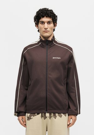 Man wearing a brown zip-up jacket with white piping and "Palm Angels" logo on chest, paired with beige pants with dark abstract print.