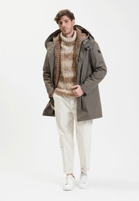 Parka impermeabile khaki con interno a quadri, abbinata a un maglione beige a trecce, pantaloni di velluto a coste color beige e sneakers bianche.