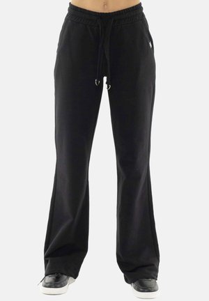 Pantalon de jogging noir à jambes larges avec taille à cordon, porté avec des baskets noires et blanches sur une personne debout.