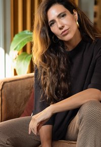 Zwarte, korte mouwen top; geblokte, bruine broek met een visgraatpatroon; lang, golvend bruin haar; gouden oorringen; gezeten op een bank.