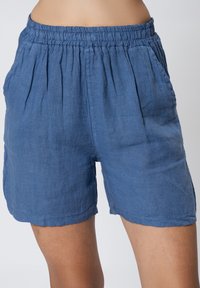 Pantaloni corti in lino blu con vita elasticizzata, tasche laterali e frontale plissettato. Il tessuto ha una consistenza morbida e una vestibilità comoda.