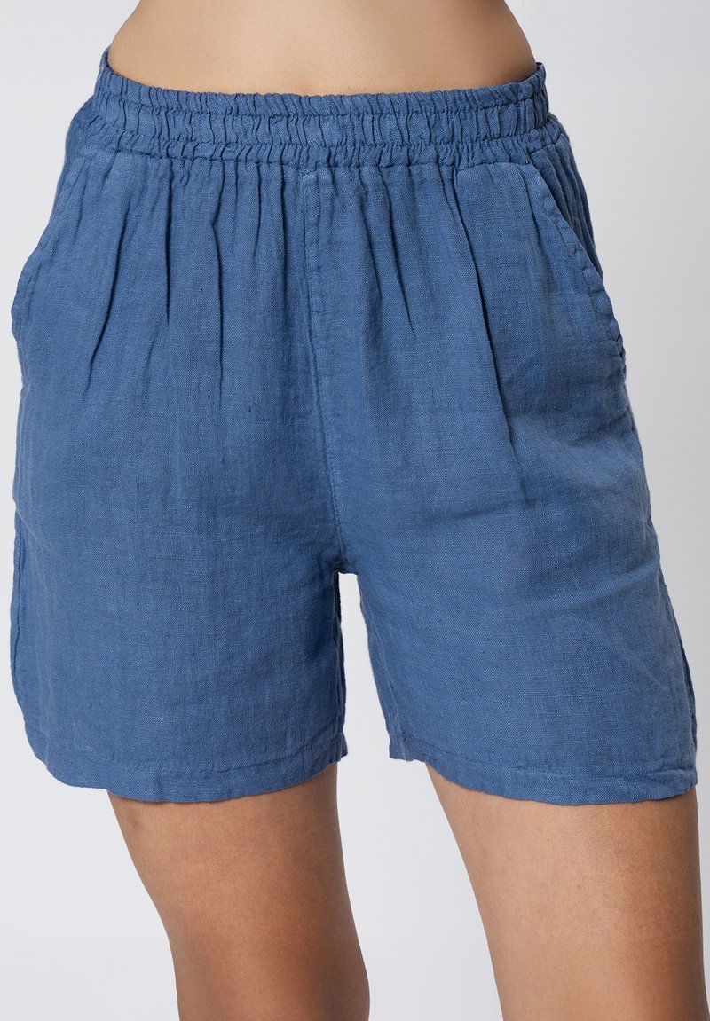 Pantaloni corti in lino blu con vita elasticizzata, tasche laterali e frontale plissettato. Il tessuto ha una consistenza morbida e una vestibilità comoda.