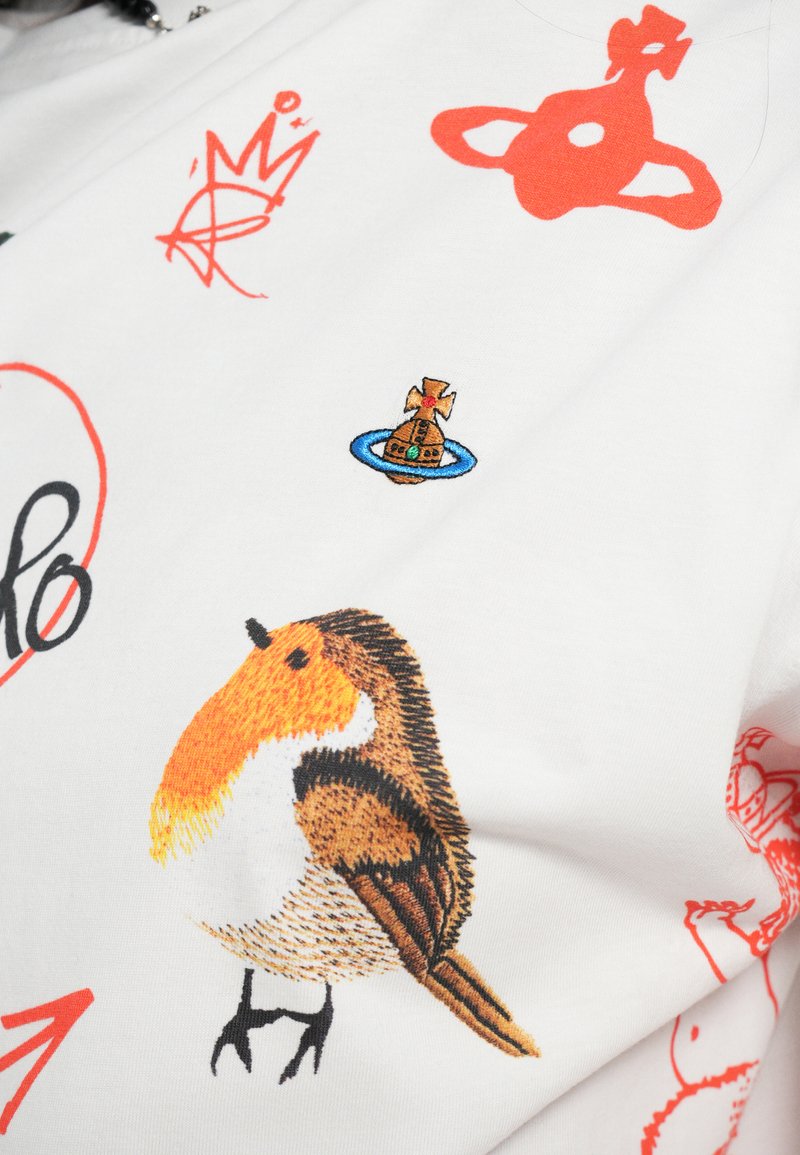 Weißes Stoffhemd mit bunten gestickten Designs, darunter ein Vogel mit orangefarbener Kappe, ein Boot mit einem Kreuz und abstrakten Formen.