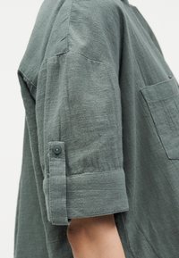 Λεπτομέρεια μανικιού γκρι-πράσινου πουκαμίσου με υφή, με γυρισμένο μανίκι που συγκρατείται από λουράκι με κουμπί και εμφανές τσέπη στο στήθος σε ανοιχτό φόντο.