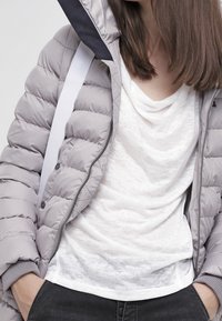Giacca imbottita grigia chiara con cappuccio nero e fascette bianche, indossata sopra una t-shirt bianca larga. Il materiale sembra morbido con una texture sottile.