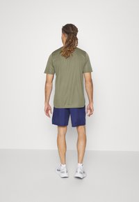 Olive green athletic t-shirt with short sleeves, paired with blue shorts. The person wears white sports shoes. Simple, functional design.