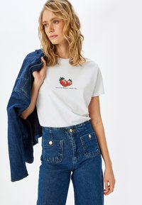 Hvid bomulds t-shirt med jordbær grafik og tekst, parret med blå højtaljede denim jeans med guldknapper og forlommer.