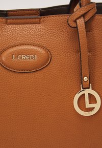 Sac fourre-tout en cuir brun avec texture granuleuse, un patch oval avec logo et un pendentif doré au design circulaire portant le nom de la marque.