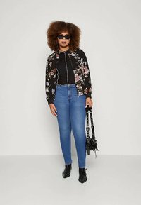 Mulher com cabelo encaracolado a usar óculos de sol, jaqueta preta floral, top preto, jeans azuis, botas pretas até ao tornozelo, segurando uma mala preta.