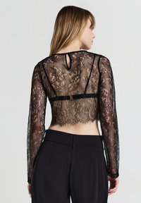 Frau, die ein schwarzes Spitzen-Crop-Top mit langen Ärmeln und schwarze hoch taillierte Hosen trägt, von hinten vor einem schlichten Hintergrund gezeigt.