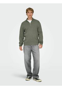 Zielony sweter na zamek z prążkowanymi mankietami, w połączeniu z jasnoszarymi jeansami i czarnymi niskimi trampkami, na neutralnym tle.