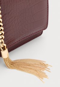 Primo piano dell'angolo di una borsa in pelle bordeaux con texture, dettagli della frangia in catena dorata su sfondo chiaro.