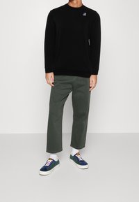 Schwarzer Sweatshirt mit Rundhalsausschnitt, gerippten Bündchen und Saum, kombiniert mit lockeren olivgrünen Hosen. Das Model trägt blaue und grüne Sneakers mit weißen Socken.