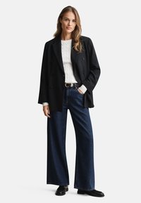 Schwarzer doppelt geknöpfter Blazer über einem weißen Kabelstrickpullover, kombiniert mit weit geschnittenen, dunkelblauen Jeans und schwarzen Schuhen. Glatte Texturen.