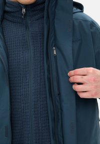 Giacca esterna blu navy con una texture liscia, dotata di zip e chiusure in velcro, sopra uno strato interno in pile blu navy con zip e texture. 