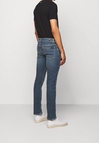 Slim-fit džíny v tmavě modré barvě, s pěti kapsami, mírně opotřebovaným vzhledem a středním pasem. Kombinováno s bílými teniskami.