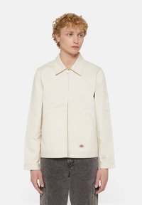 Beige cotton jacket with a collared neckline, front zipper, side pockets, and logo patch. Features a relaxed fit and long sleeves.