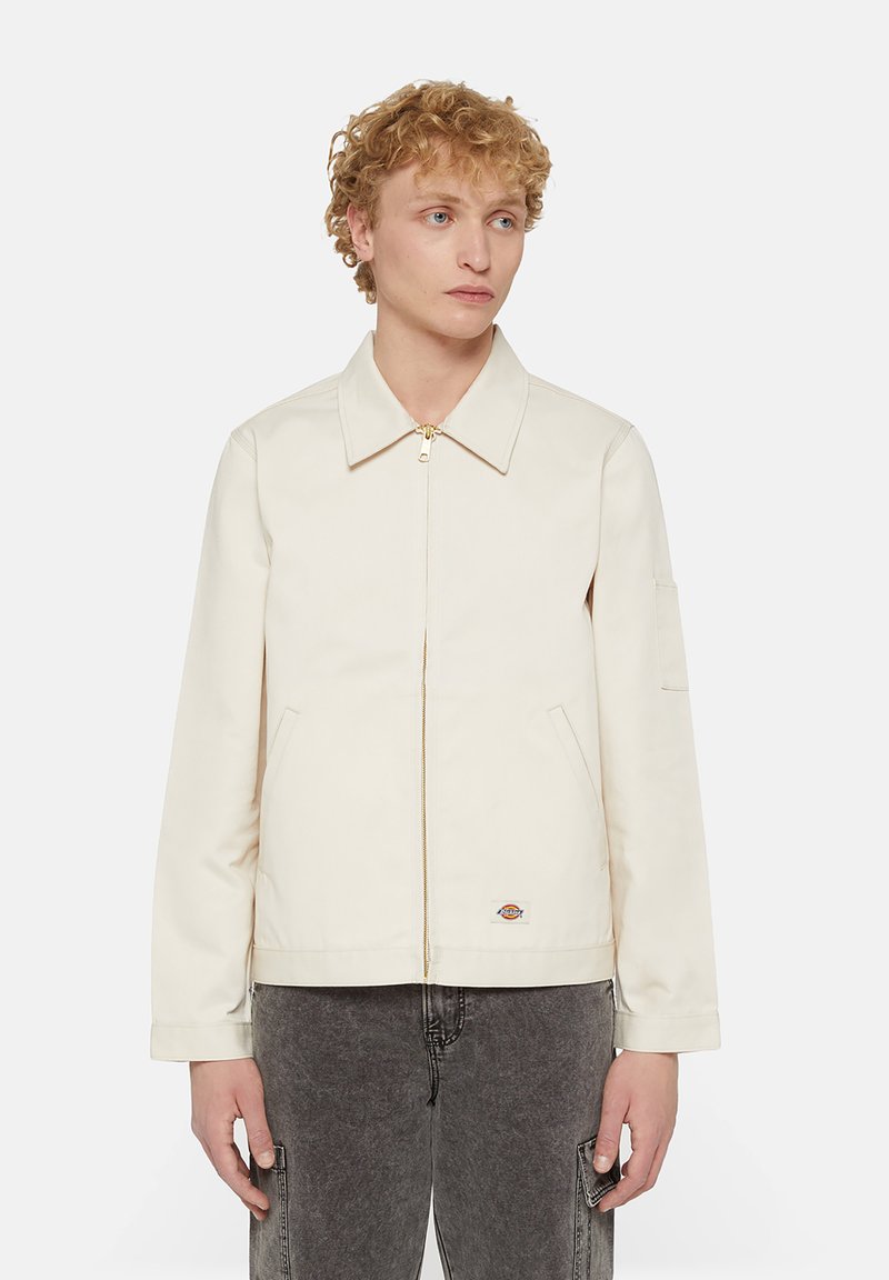 Beige cotton jacket with a collared neckline, front zipper, side pockets, and logo patch. Features a relaxed fit and long sleeves.