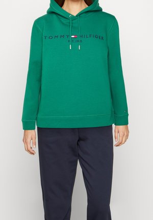 Personne portant un sweat à capuche vert Tommy Hilfiger avec logo et un pantalon bleu marine, debout sur un fond uni clair.