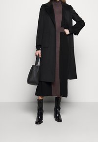 Nainen, jolla on päällä musta pitkä takki mustan ja ruskean korkeakauluksisen mekon päällä, mustat nahkasaapikkaat nilkkoihin ja hän kantaa mustaa nahkaista käsilaukkua.