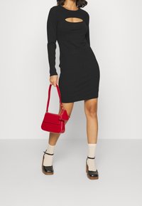 Robe noire à manches longues avec un détail découpé, associée à un sac à main rouge, des chaussettes crème et des chaussures compensées noires à talons en bois.