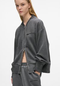 Chaqueta bomber gris con cremallera, hecha de algodón suave, con puños acanalados y un logo de "KARL LAGERFELD JEANS" en el frente.