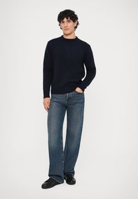 Maglione navy con lavorazione a maglia testurizzata e colletto a girocollo, abbinato a jeans a gamba larga di un blu chiaro e sneakers nere. Il modello è in piedi con una mano in tasca.