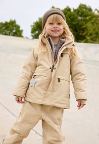 Beige Winterjacke mit Kapuze, zwei Fronttaschen und einem Sternakzent, kombiniert mit passenden beigen Hosen. Hat eine weiche, strukturierte Optik.