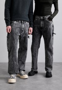 To personer har overdimensionerede, falmede sorte denimbukser med flere lommer. De parrer dem med kontrasterende fodtøj: en lys og en mørk.
