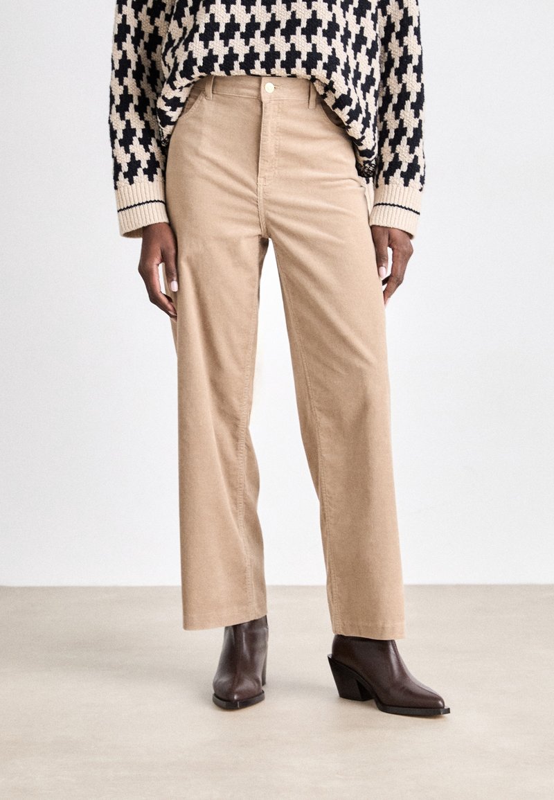 Personne portant un pantalon large beige taille haute, un pull à motif pied-de-poule noir et blanc, et des bottines à talons brun foncé se tenant sur un sol neutre.