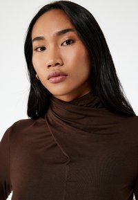 Top marrón ajustado con un cuello drapeado alto. Tejido suave y liso con costuras mínimas, que muestra un diseño limpio y sencillo.