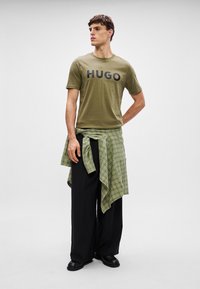 Olivová zelená tričko s potlačou "HUGO" v čiernej farbe, spojené s voľnými čiernymi nohavicami a zeleno-čierne kárované košeľou zviazanou okolo pása.