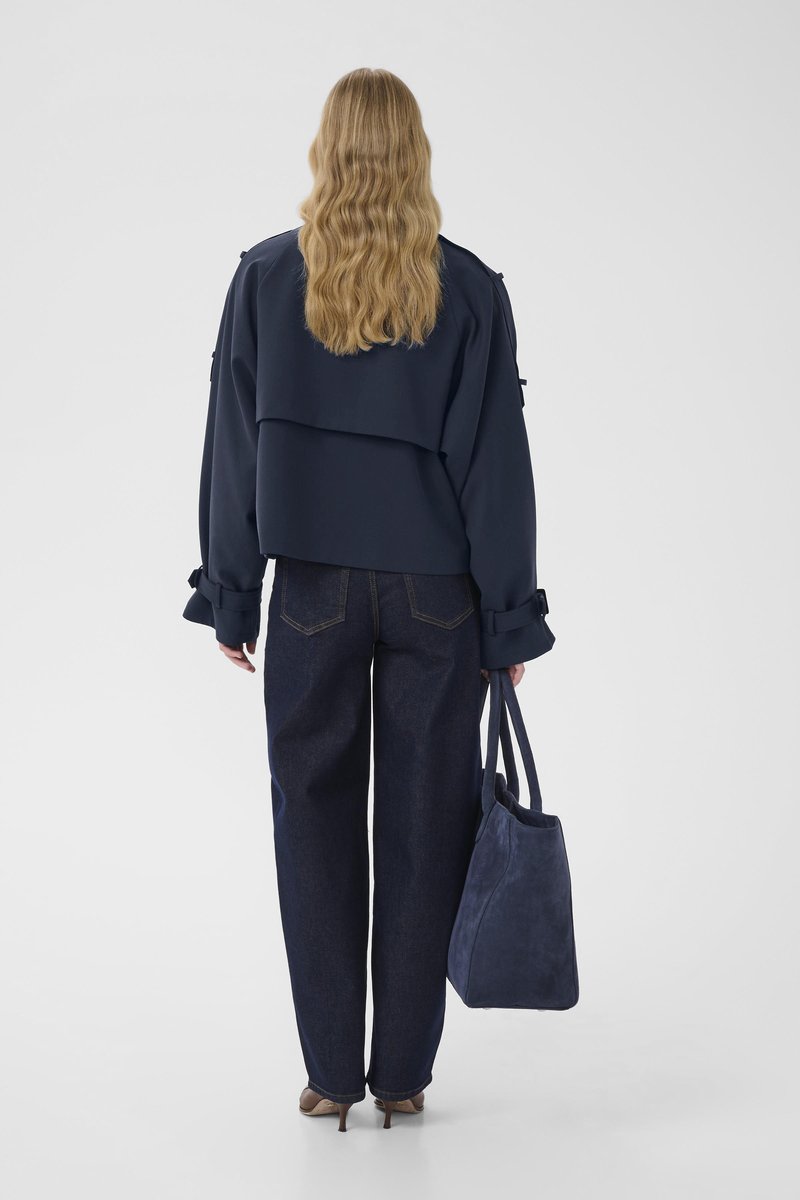 Veste courte bleu marine à dos superposé, associée à un jean denim foncé à jambes larges. Tient un sac fourre-tout en daim bleu marine. Chaussures à talons de couleur neutre.