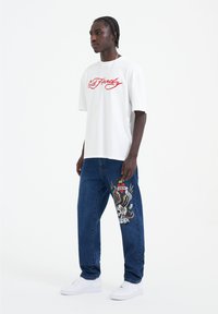 Camiseta blanca de manga corta con "Ed Hardy" en rojo, combinada con jeans de mezclilla azul que tienen un gráfico bordado colorido en la pierna izquierda.