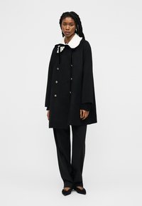 Frau trägt einen schwarzen, doppelreihigen Mantel mit weißem Pelzkragen, schwarze Hose und schwarze spitze Schuhe, steht vor einem einfarbigen Hintergrund.