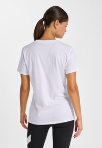 Femme aux cheveux bruns attachés en queue de cheval, portant un t-shirt blanc uni à manches courtes et des leggings noirs, vue de dos sur un fond clair uni.