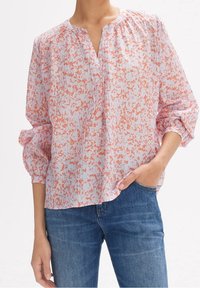 Blusa a maniche lunghe di un delicato rosa con un motivo aranciato a pois. Presenta un collo a V, dettagli arricciati e maniche larghe. Indossata sopra un paio di jeans blu.