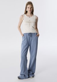 Crème tanktop met decoratief ontwerp en knopen; losse blauwe sweatpants met trekkoord en studs langs de naden.
