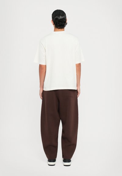 Weißes Baumwoll-T-Shirt mit kurzen Ärmeln, kombiniert mit locker sitzenden braunen Hosen. Die Hosen haben eine Gesäßtasche und einen strukturierten Stoff.
