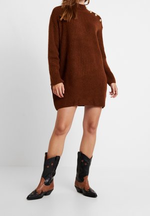 Femme portant une robe-pull en tricot marron avec des boutons dorés sur l'épaule et des bottes de cowboy en cuir noir et marron avec des détails en forme d'étoile.