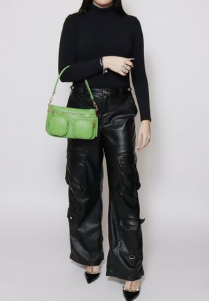 Femme portant un col roulé noir et un pantalon cargo en cuir, tenant un sac à main vert avec deux poches avant sur un bras.