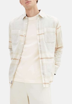 Lys beige ternet skjorte med to brystlommer, knaplukning foran og lange ærmer. Bæres over en cremefarvet t-shirt, parret med lyse bukser.