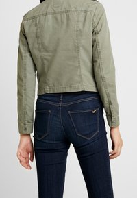 Olivgrön kort jacka med knäppte manschetter och enkla sömmar, tillsammans med mörkblå jeans som har bakfickor och sömnadsdetaljer.