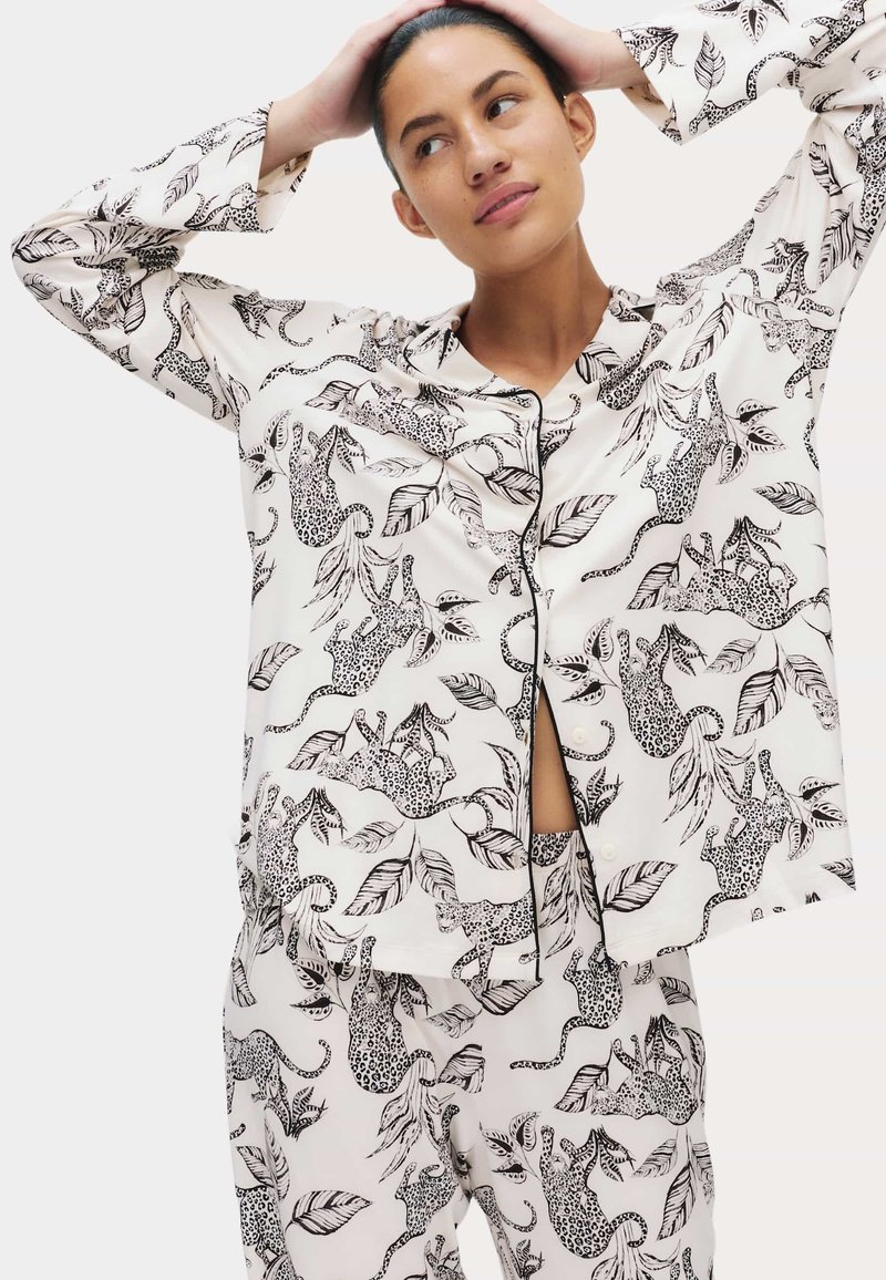 Kvinde iført hvidt pyjamasæt med sort leopard- og bladprint, der kigger til siden med hænderne på hovedet.