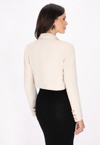 Gekropte beige jas met lange mouwen, voorzien van parelknopenaccenten op de manchetten, gecombineerd met een aansluitende zwarte rok. Soepele stoftextuur.