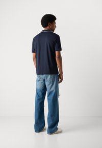 Ναυτικό πόλο μπλουζάκι με κλασικό γιακά, συνδυασμένο με ανοιχτό μπλε φθαρμένα φαρδιά τζιν και λευκά αθλητικά παπούτσια, θέα από πίσω.