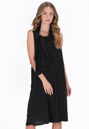 Kobieta o falowanych włosach, ubrana w czarną, luźną sukienkę bez rękawów z drapowanym szalowym detalem, stojąca na białym tle.