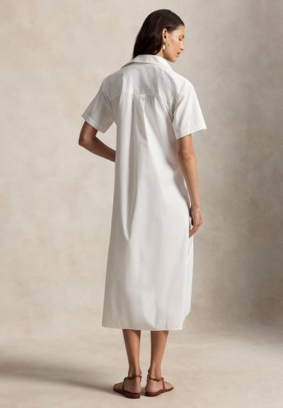 Femme portant une robe-chemise blanche à manches courtes et longueur midi avec col, debout de dos, chaussée de sandales plates marron.