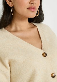 Cardigan en tricot beige avec un décolleté en V profond et trois boutons marron, en tissu doux et texturé. Comprend de grandes boucles d'oreilles en or.